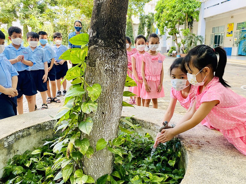 Hình ảnh cô giáo đang chỉ dẫn các em nhỏ rửa tay dưới sân trường.